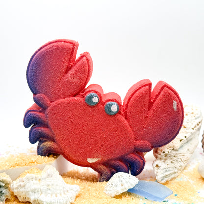 Handmade red crab bath bomb in sand displayed with shells and sand. 