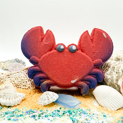 Hand made red crab bath bomb with blue pinchers displayed on a beach themed background. 