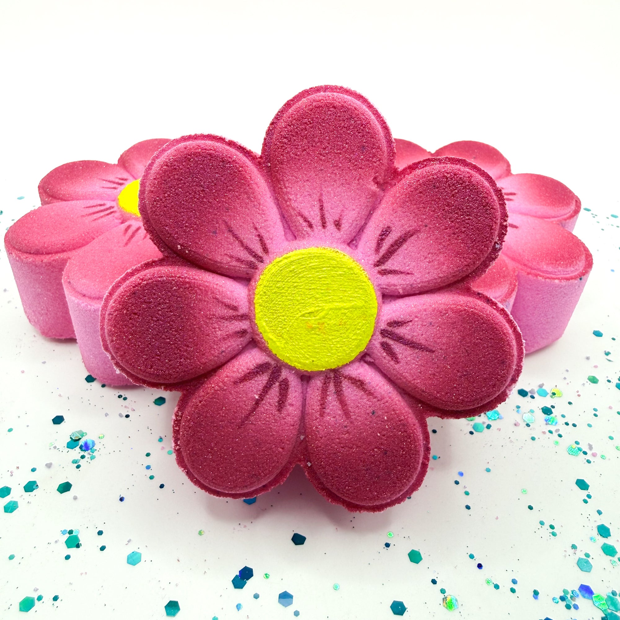 Hand made pink daisy bath bomb with a yellow center on a white background with colorful confetti.