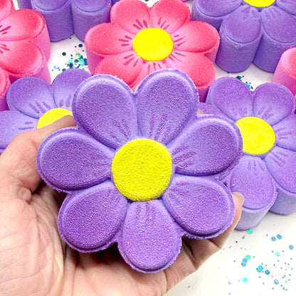 Hand painted purple daisy bath bomb with a yellow center held by a hand, surrounded by similar objects on a white background.