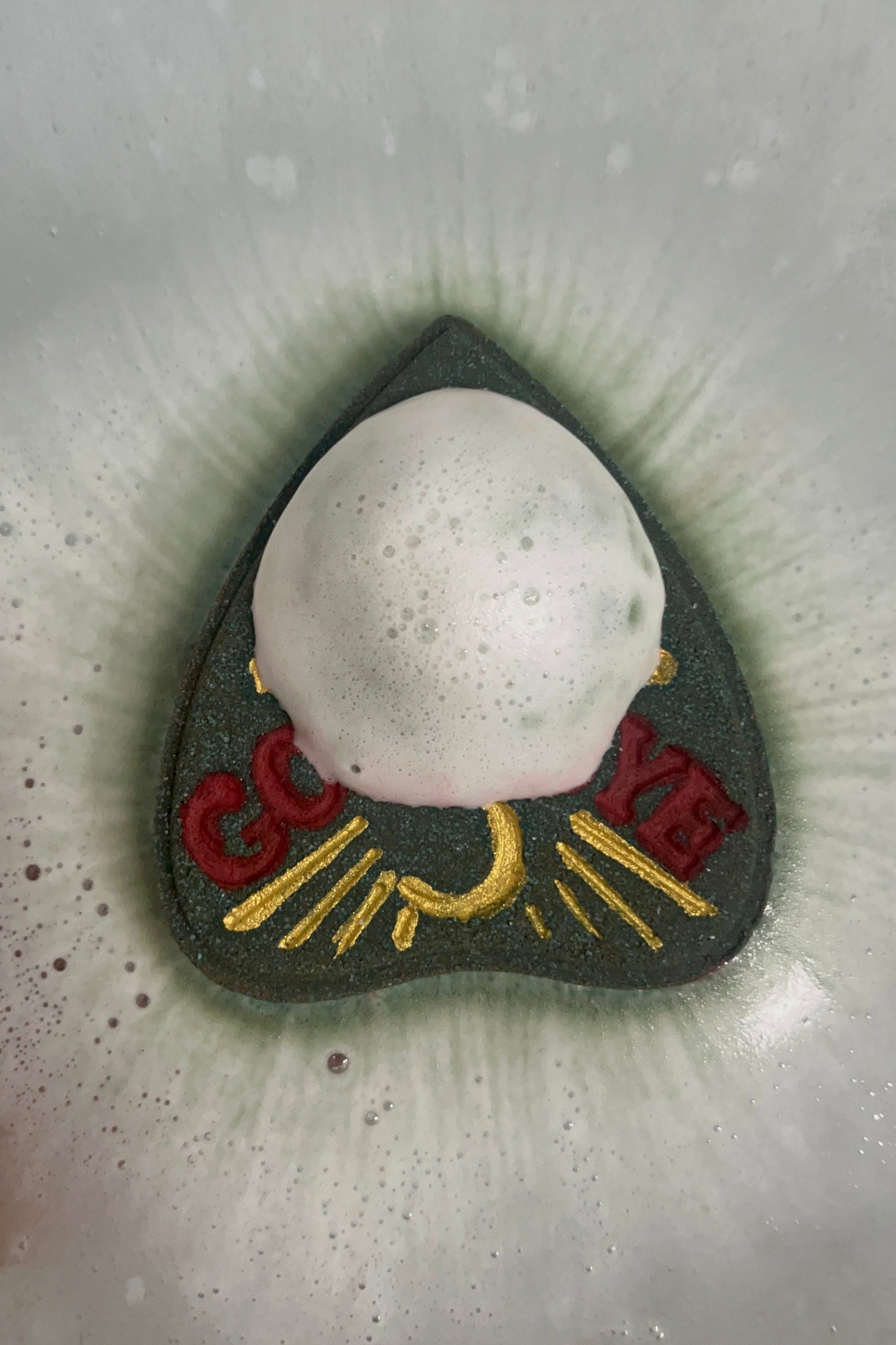 A dark colored planchette shaped bath bomb from Rusted Acre Soap Company in water surrounded by fizz and foam.