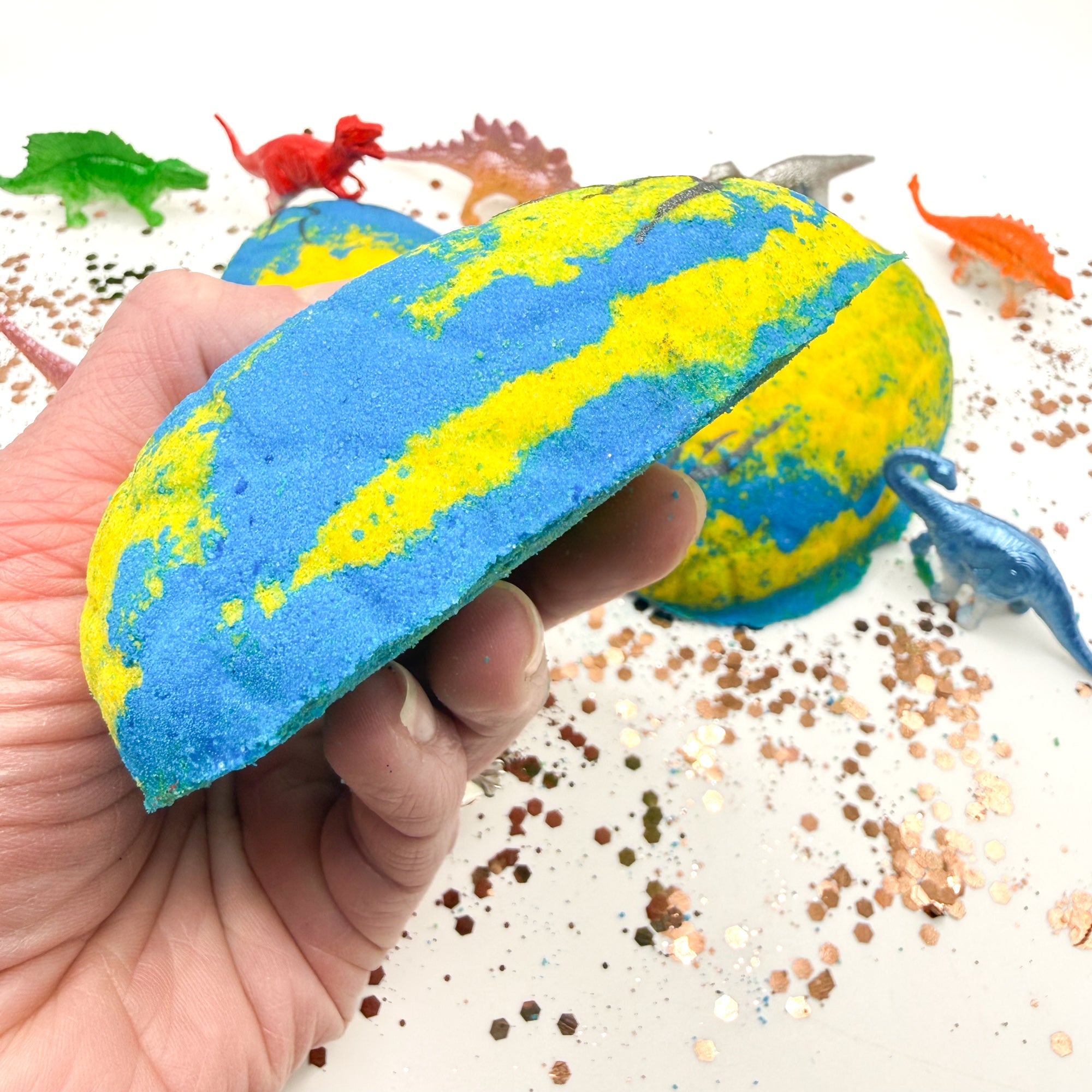Yellow and Blue dinosaur egg bath bomb held by a hand with bath toys in the background made by Rusted Acre Soap Co.