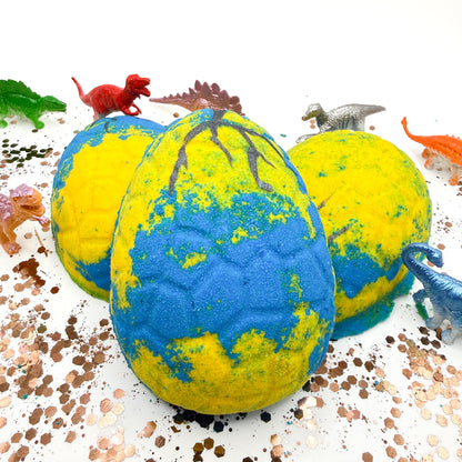 Dinosaur egg bath bombs with blue and yellow pattern, surrounded by small dinosaur figurines that are inside the bath bomb on a white background. 