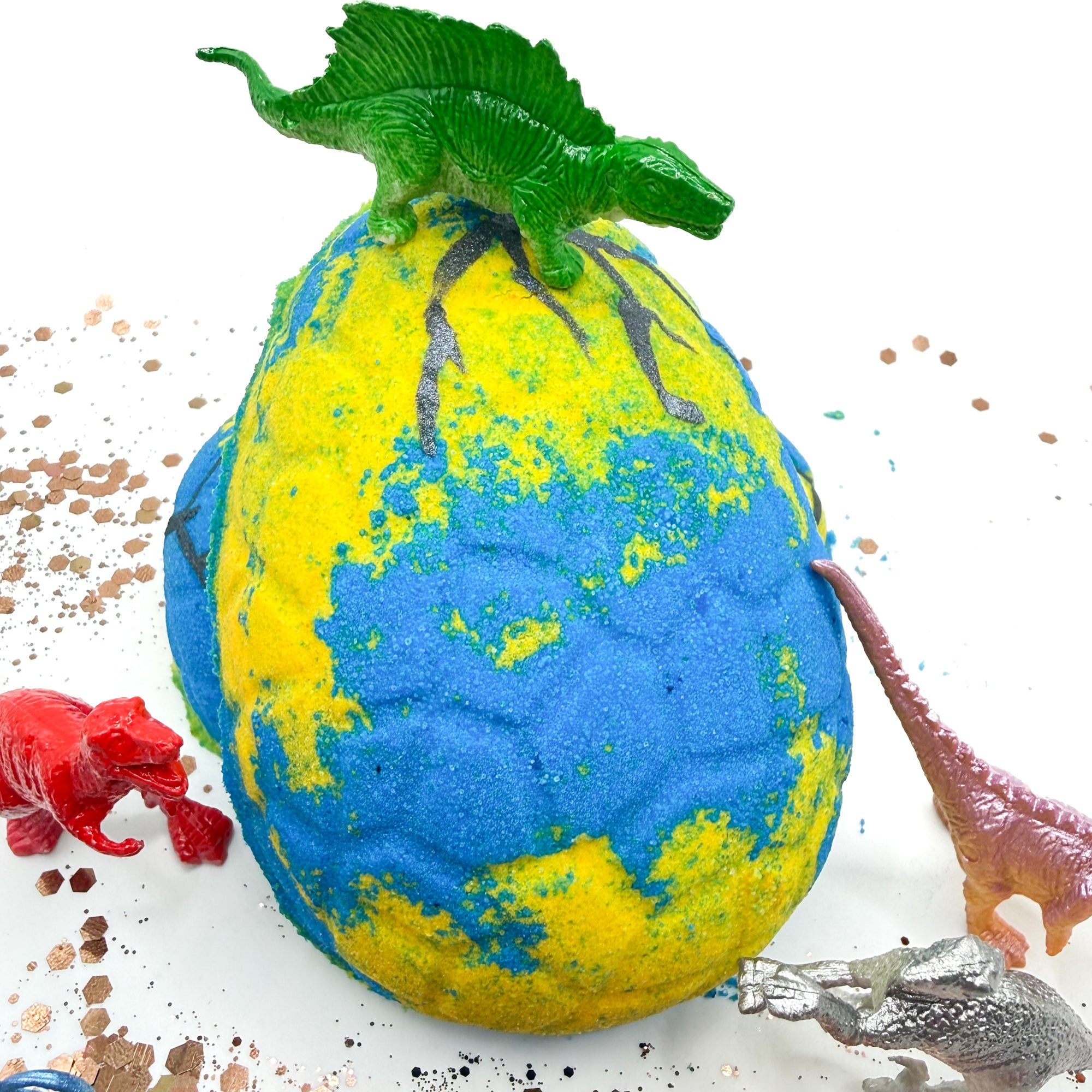 Blue and yellow dinosaur egg bath bomb with a dinosaur on top, surrounded by small toy dinosaurs surrounding it with a background.