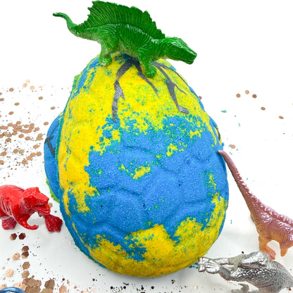 Blue and yellow dinosaur egg bath bomb with a dinosaur on top, surrounded by small toy dinosaurs surrounding it with a background.
