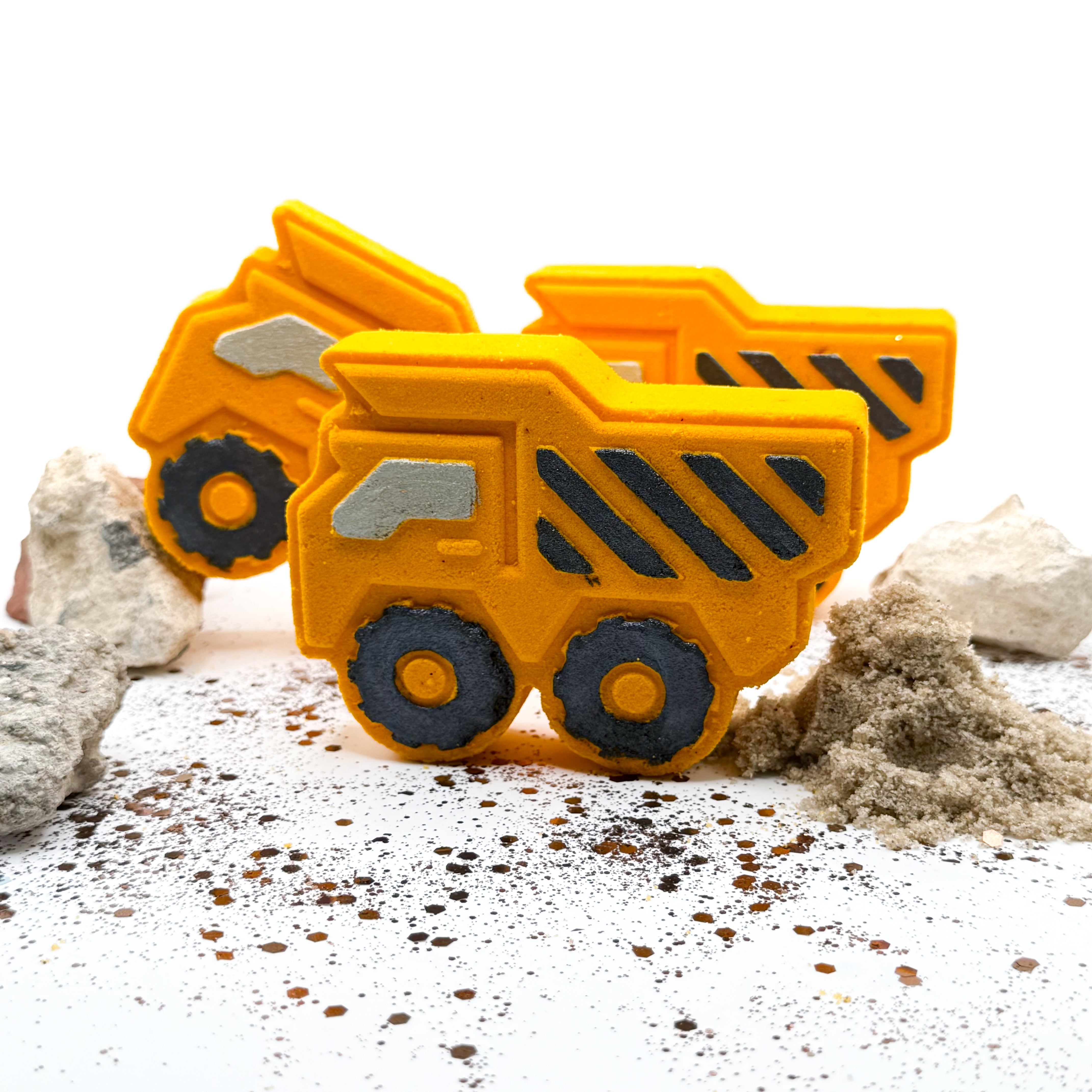 Two bright yellow dump truck bath bombs hand painted with black and gray details, set on a white surface with sand, rocks, and scattered crumbs.