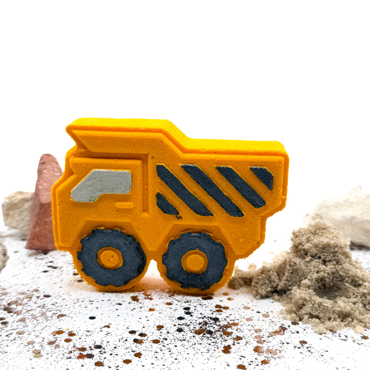 Yellow dump trucks bath bombs made by Rusted Acre soap Co. Black hand painted stripes and other details, standing up with rocks behind it.