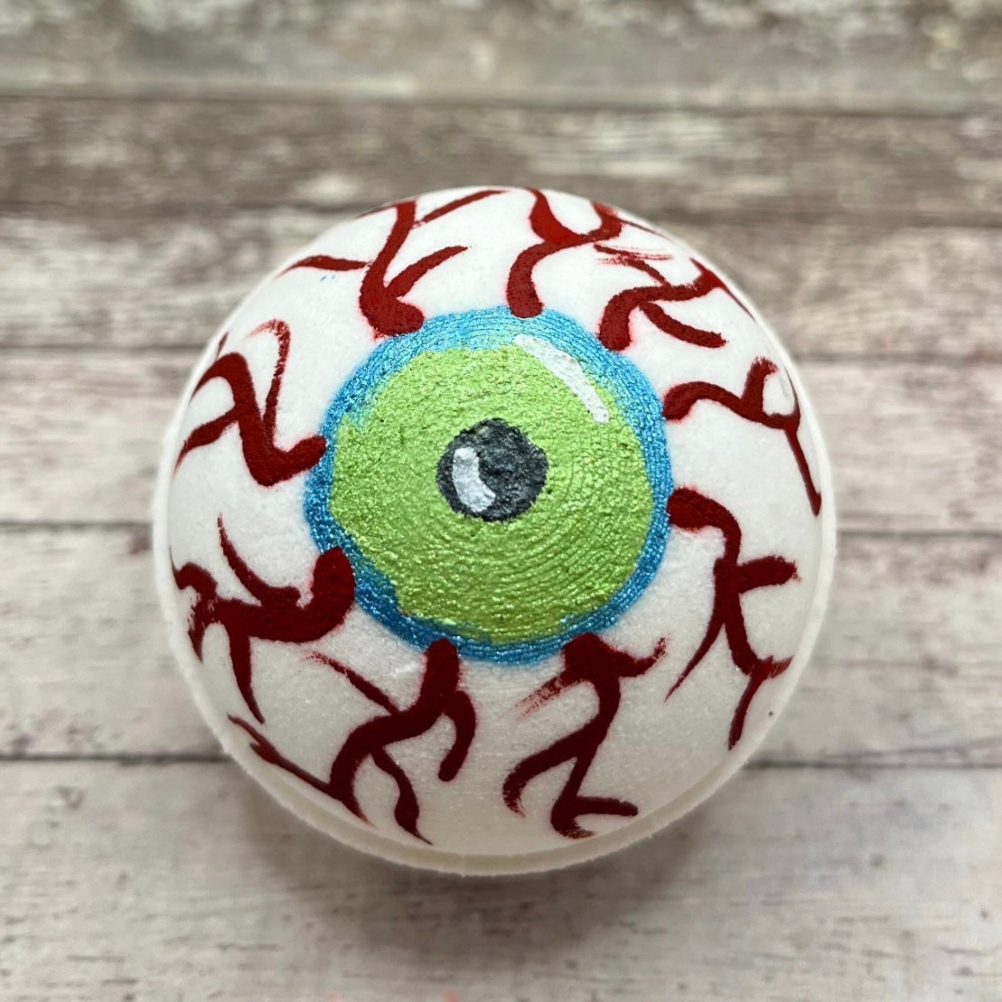 Hand-painted eyeball bath bomb with green iris, blue outline, and red vein details on a white background hand made by Rusted Acre Soap Co.
