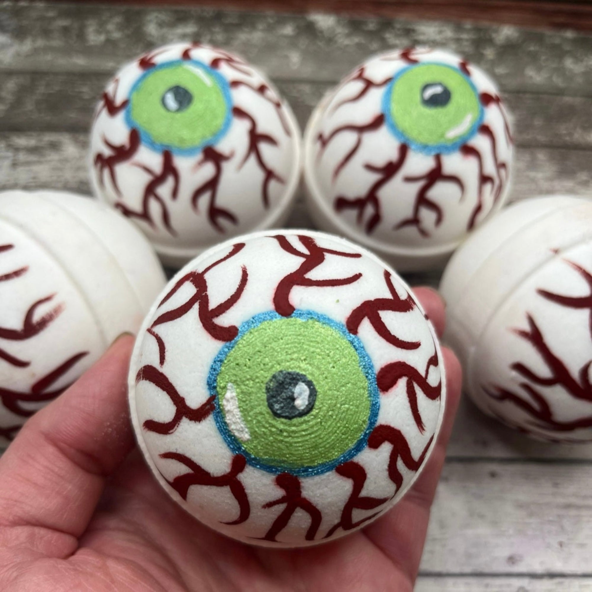 Hand holding a round eyeball bath bomb with green iris, blue outline, and red vein details, with several matching bath bombs in the background made by Rusted Acre Soap Co.
