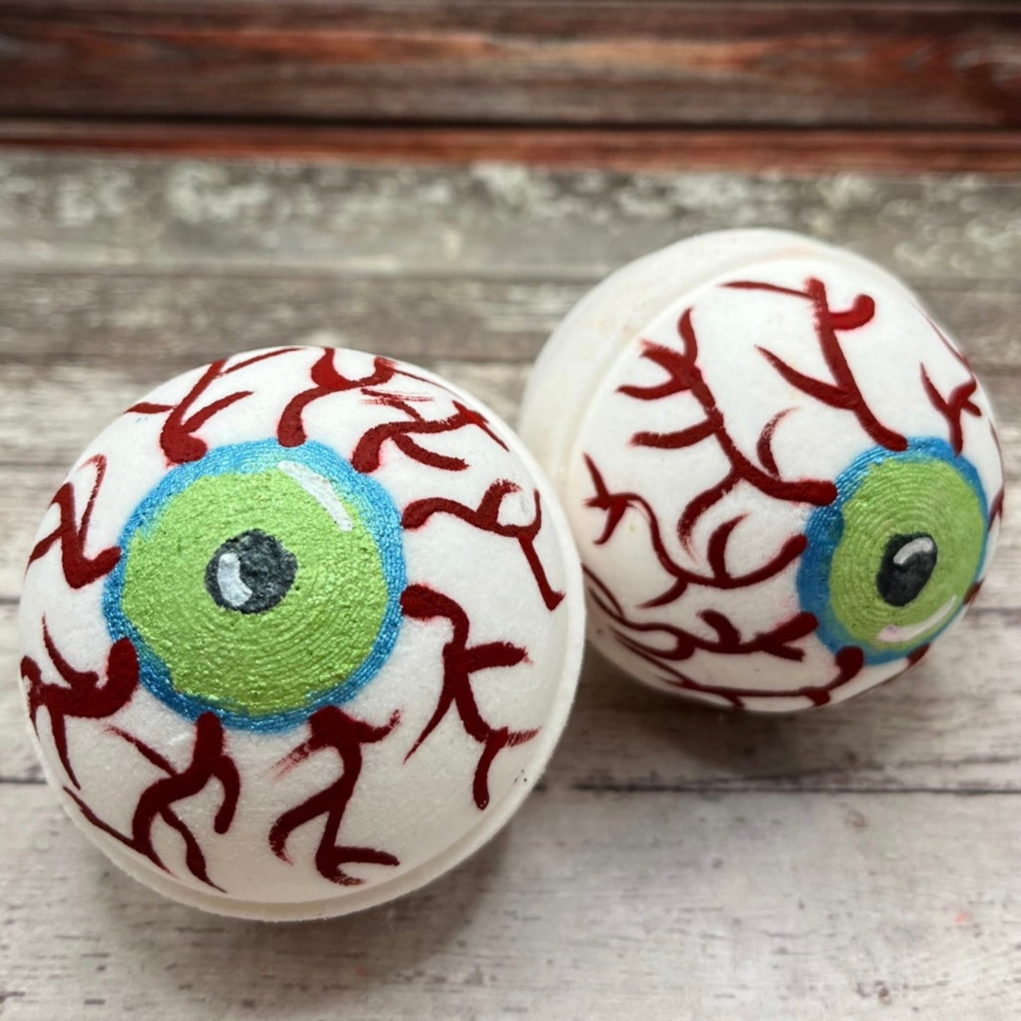 Two eyeball bath bombs with green irises, blue outlines, and red vein details resting on a wooden surface made b Rusted Acre Soap Co.
