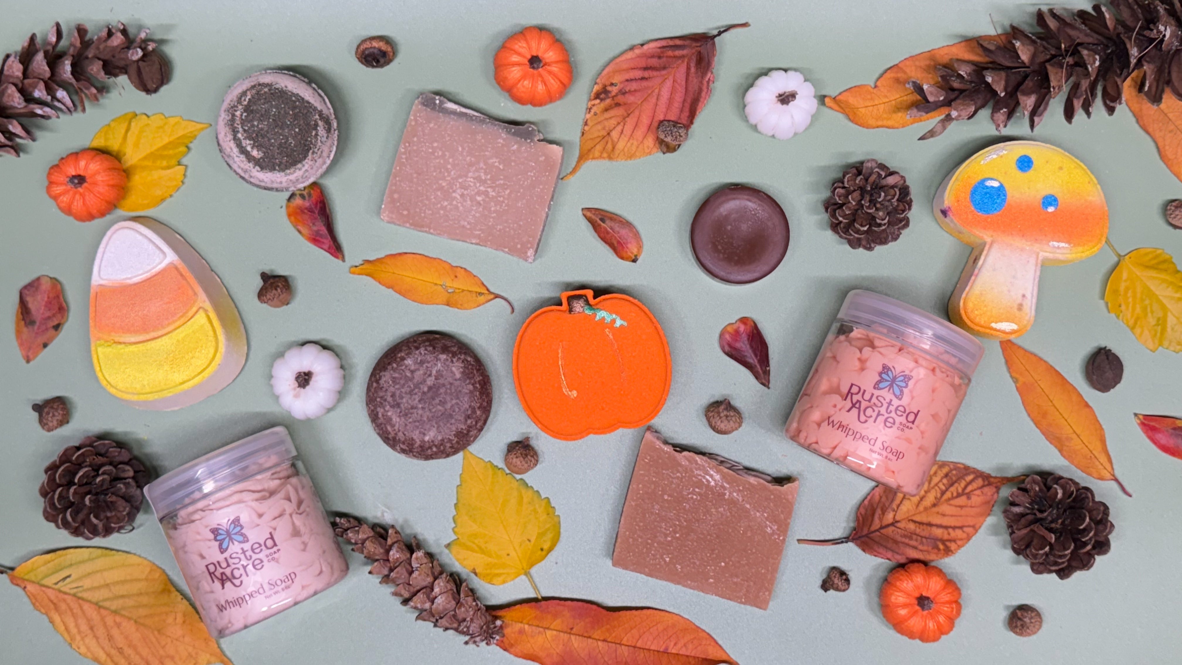 Assorted Fall themed products including handmade soap, whipped soap, hand painted bath bombs and mini soap pumpkins surrounded by fall leaves and acorns. 