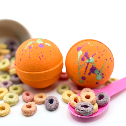 Two orange fruity cereal bath bomb with green and pink specks on top. Pictured on a white  background with cereal loops scattered about 