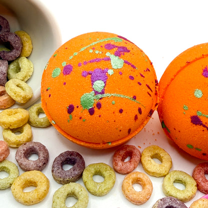 One orange fruity cereal bath bomb with green and pink specks on top. Pictured on a white  background with cereal loops scattered about 
