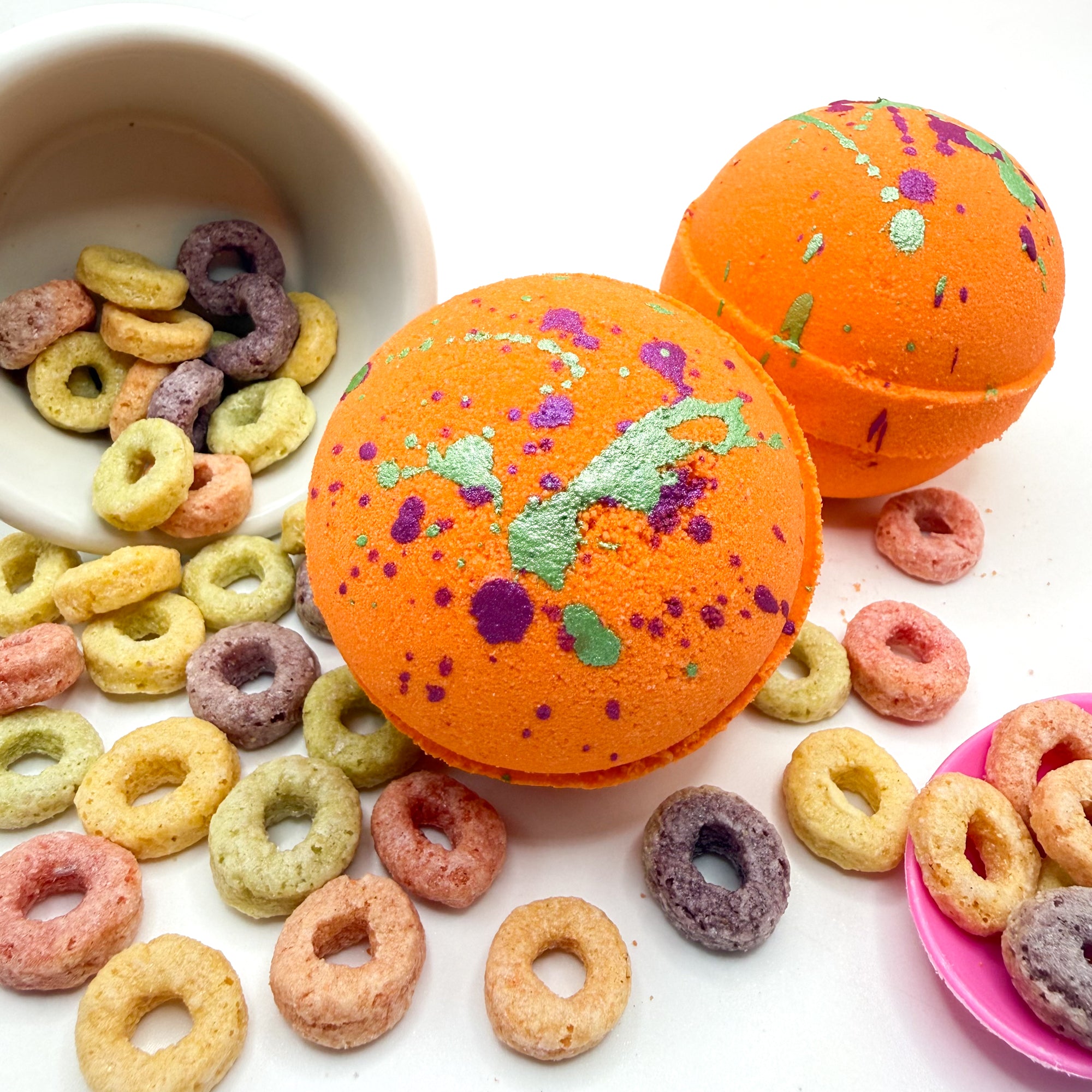 Two orange fruity cereal bath bomb with hand painted green and pink specks on top. Pictured on a white  background with cereal loops scattered about 