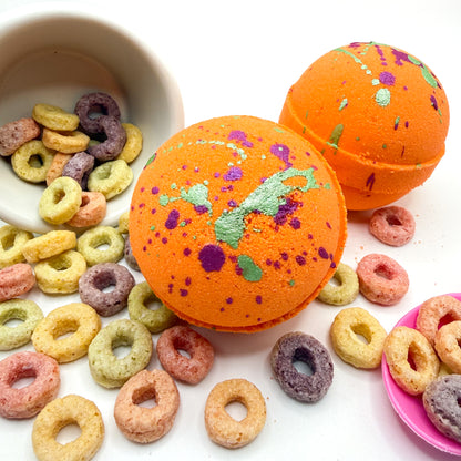 Two orange fruity cereal bath bomb with hand painted green and pink specks on top. Pictured on a white  background with cereal loops scattered about 