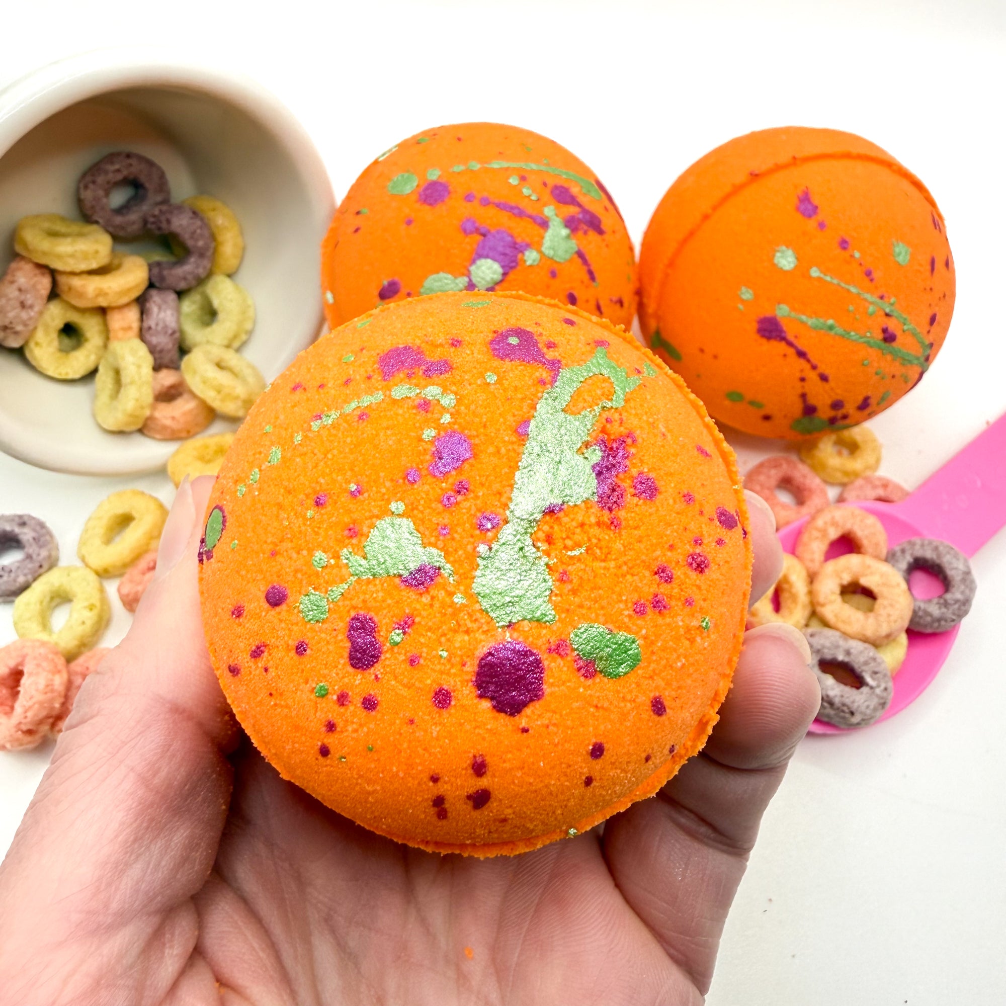 Orange fruity cereal bath bomb with hand painted pink and green splashes held by a hand, with cereal in the background.