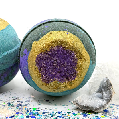 Two green, blue and purple bath bombs one facing forward and the other one facing upright with purple and gold centers resembling a geode on a glittery surface with crystals. 