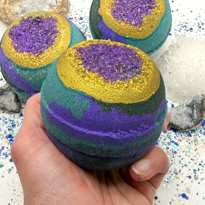 One green, blue and purple bath bomb held in a hand with purple and gold centers resembling a geode on a glittery surface with crystals.