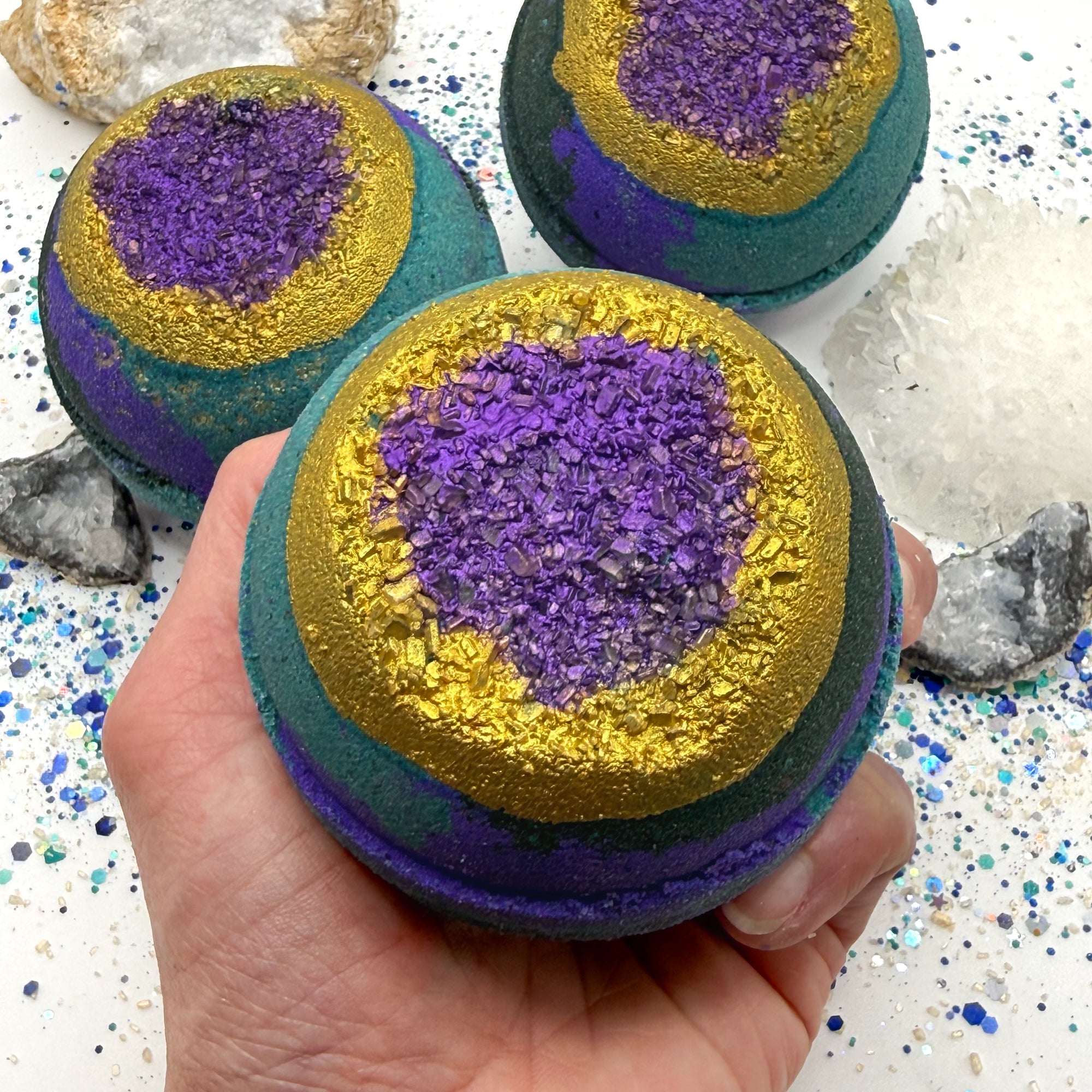 One green, blue and purple bath bomb held in a hand with purple and gold centers resembling a geode on a glittery surface with crystals.