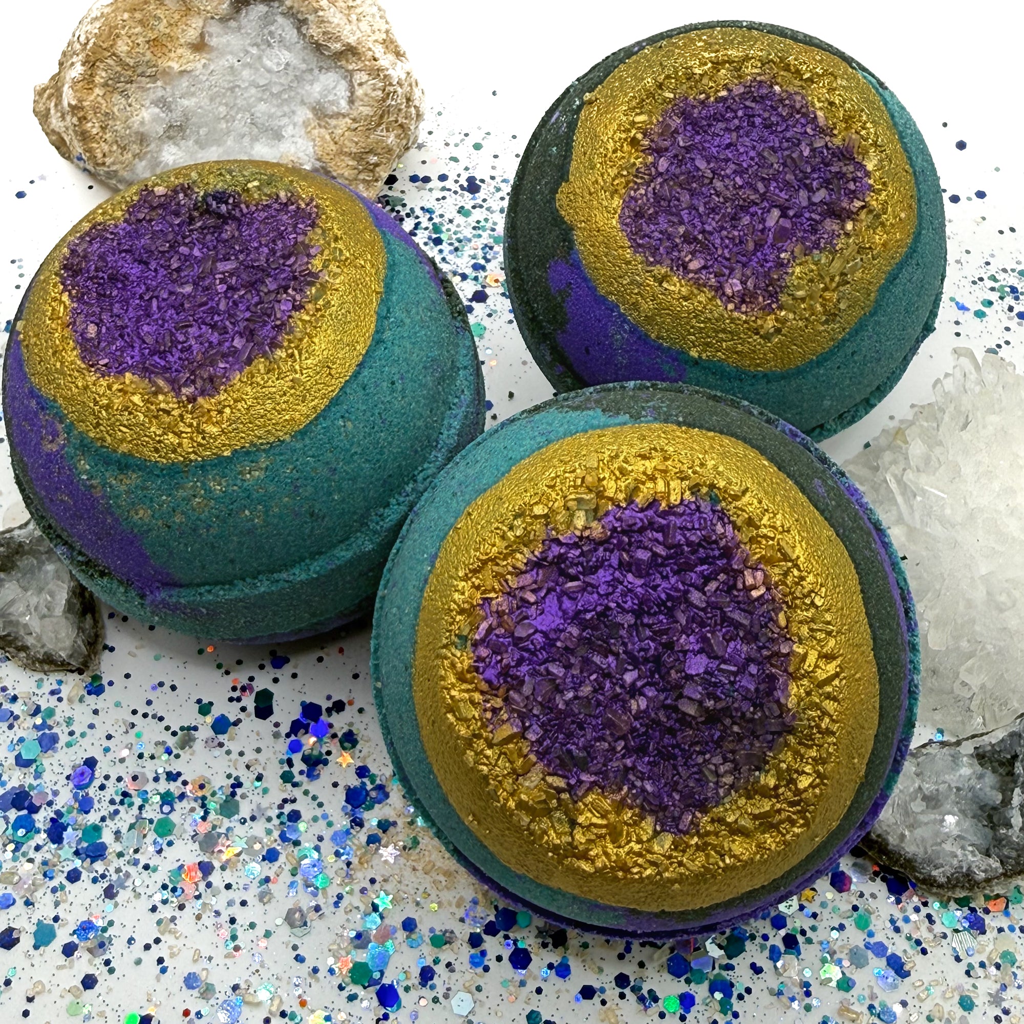 Three green, blue and purple bath bombs with purple and gold centers resembling a geode on a glittery surface with crystals.