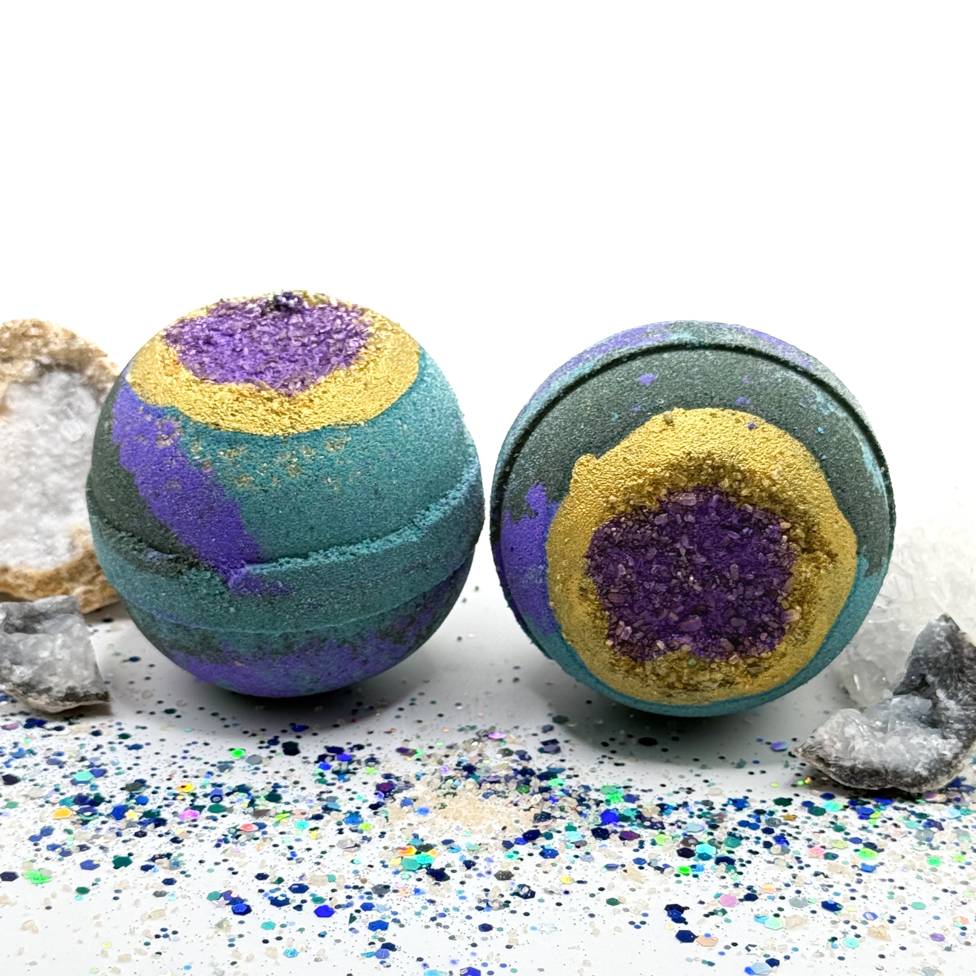 Two hand painted green, blue and purple bath bombs with purple and gold centers resembling a geode on a glittery surface with crystals.