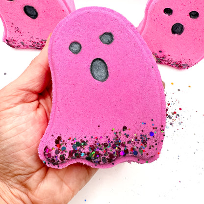Hand holding a pink ghost-shaped Halloween bath bomb from Rusted Acre Soap Co. with black eyes and colorful glitter at the base. 