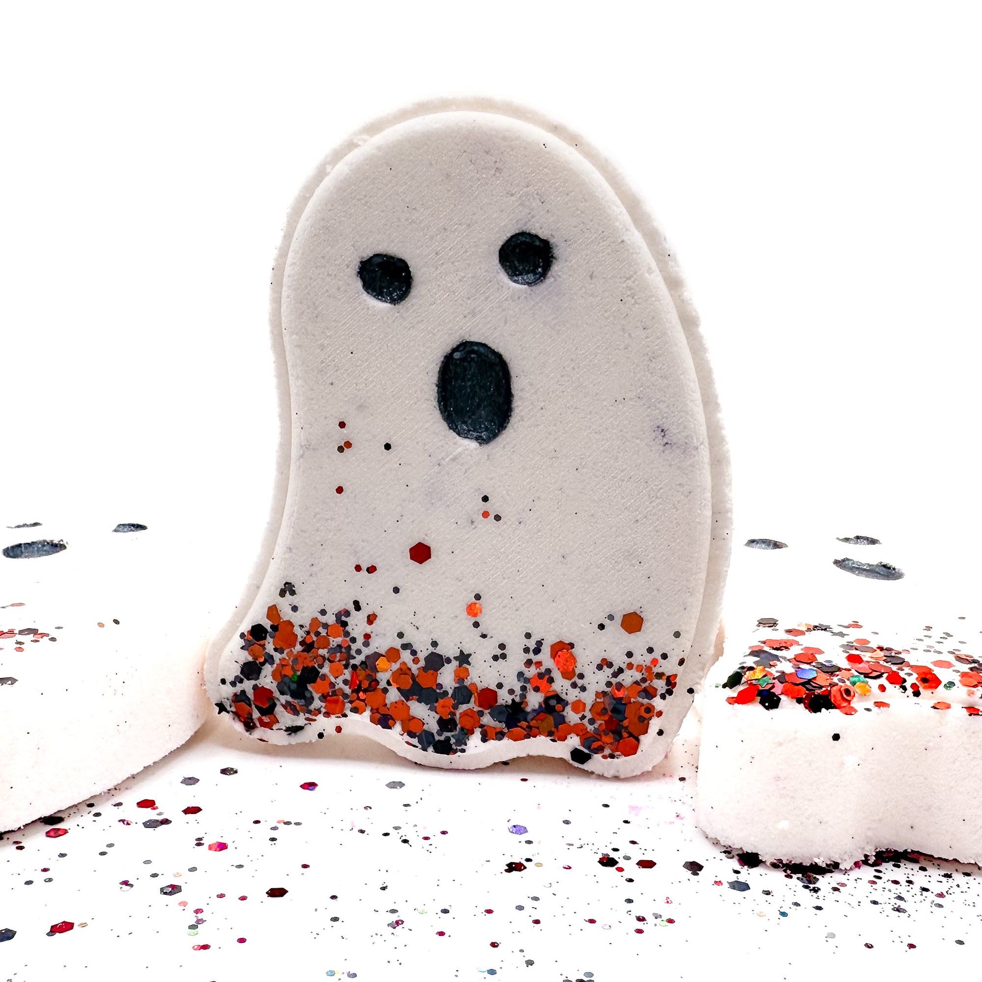 White ghost bath bomb from Rusted Acre Soap Co. standing upright with black eyes, a surprised mouth, and orange and black glitter along the base