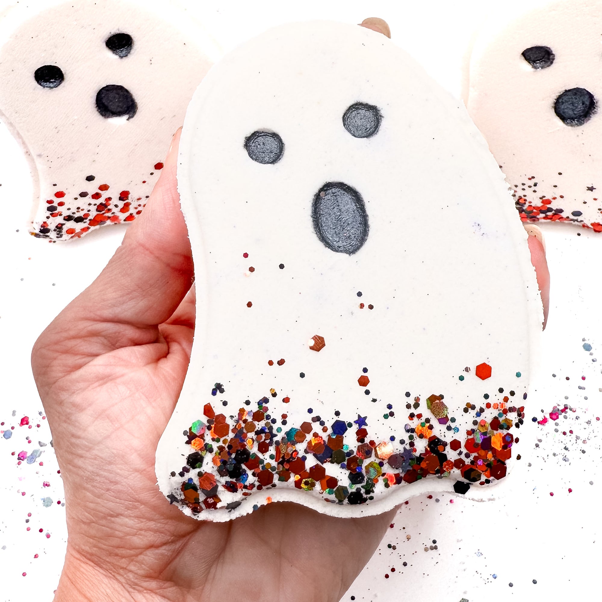 Hand holding a white ghost-shaped Halloween bath bomb from Rusted Acre Soap Co., with black eyes, a surprised mouth, and colorful glitter at the base.