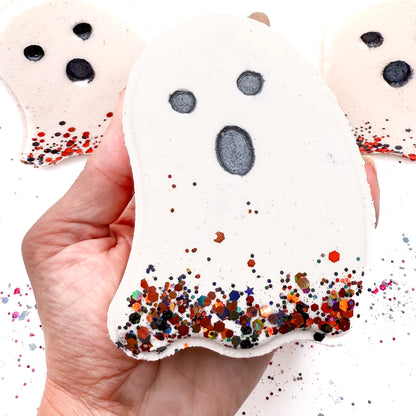 Hand holding a white ghost-shaped Halloween bath bomb from Rusted Acre Soap Co., with black eyes, a surprised mouth, and colorful glitter at the base.