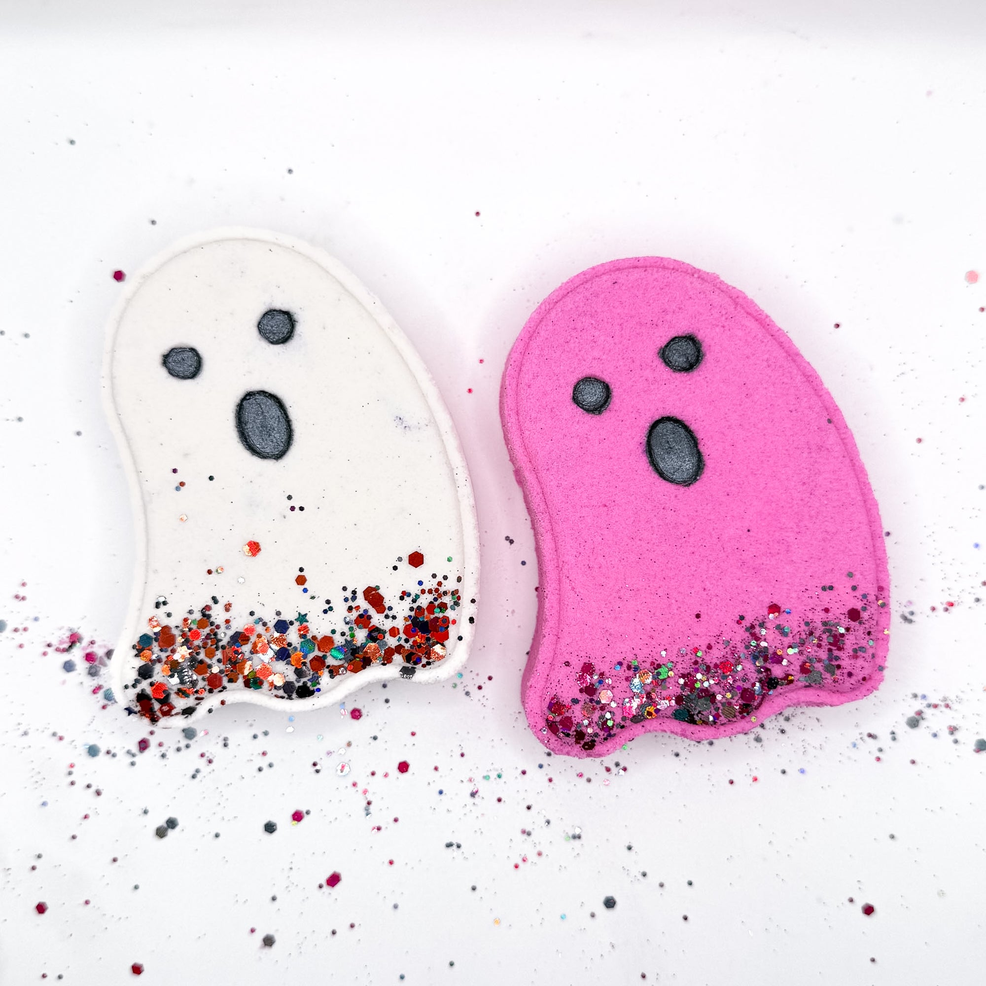 White and pink ghost-shaped Halloween bath bombs from Rusted Acre Soap Co., both with black facial features and glittery bottoms, perfect for spooky bath time fun.