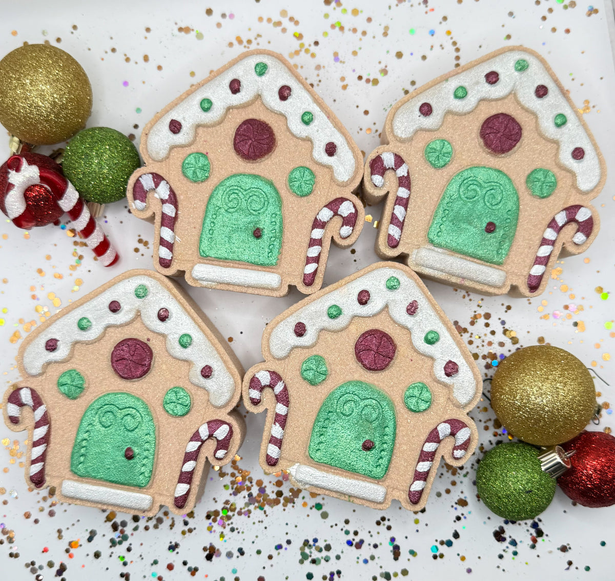 Four tan red and green hand painted gingerbread house bath bombs on a glittery gold and white background. 