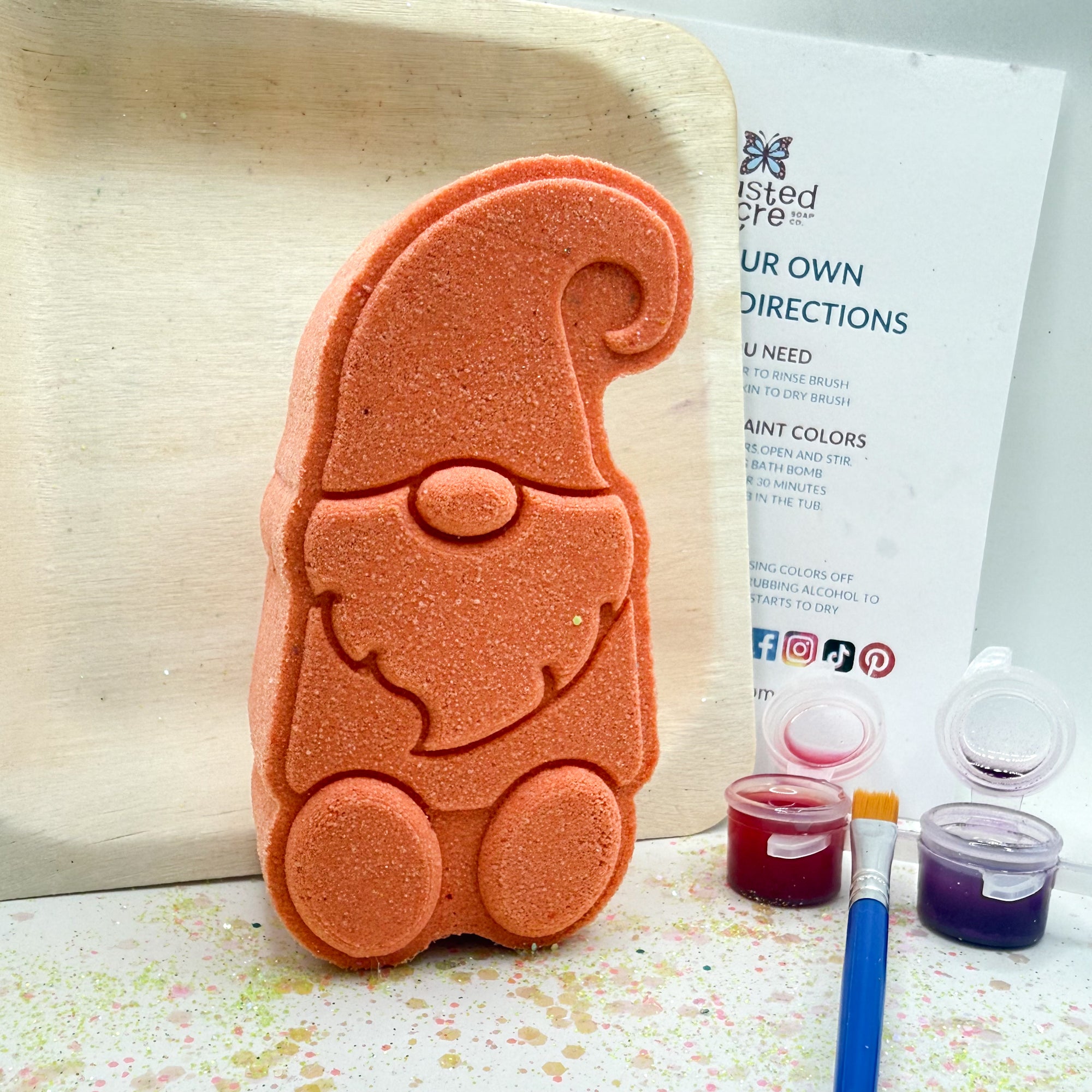 One white gnome shaped bath bomb. Displayed with the contents, a paint brush, three pots of colored mica, witch hazel and instructions.