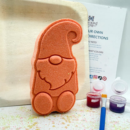 One white gnome shaped bath bomb. Displayed with the contents, a paint brush, three pots of colored mica, witch hazel and instructions.