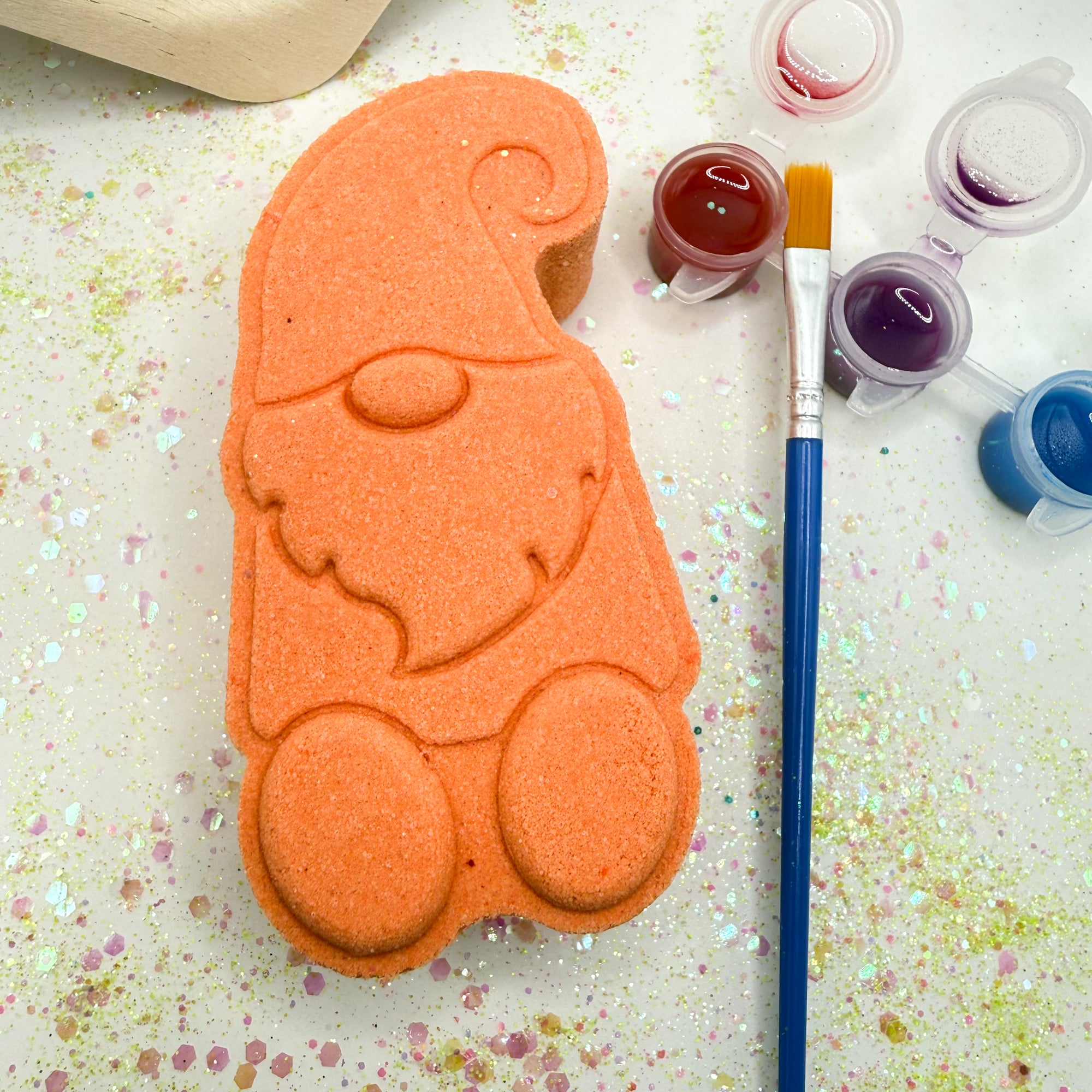 Orange gnome bath bomb with paint your own supplies, containers and a brush on a glittery surface.