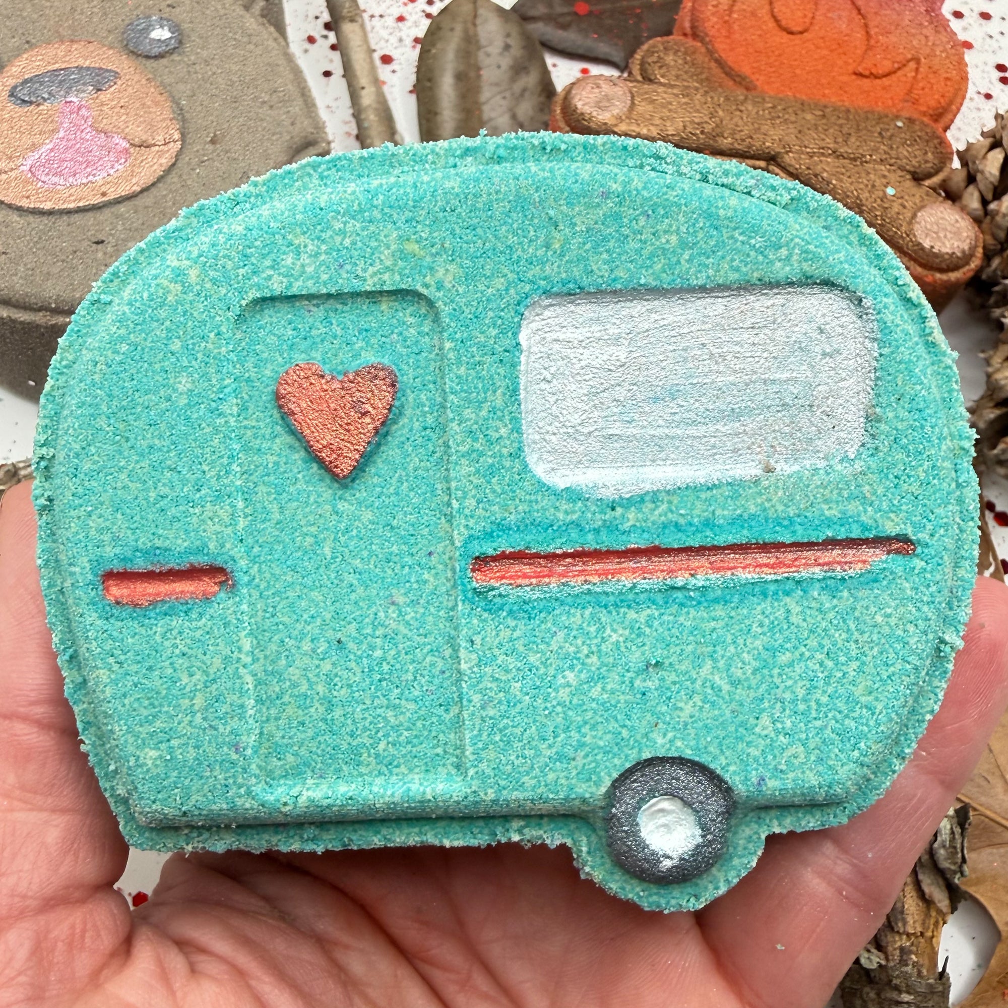 Handmade teal camper shaped bath bomb with a heart in a hand against a decorative background