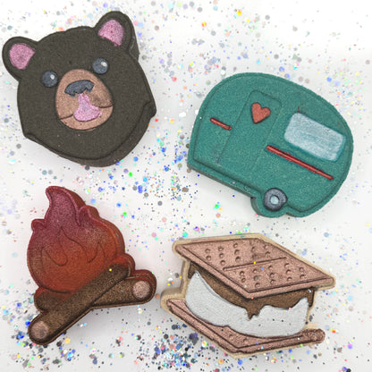 Four handmade bath bombs on a glittery background: bear, camper, campfire, and s'mores.