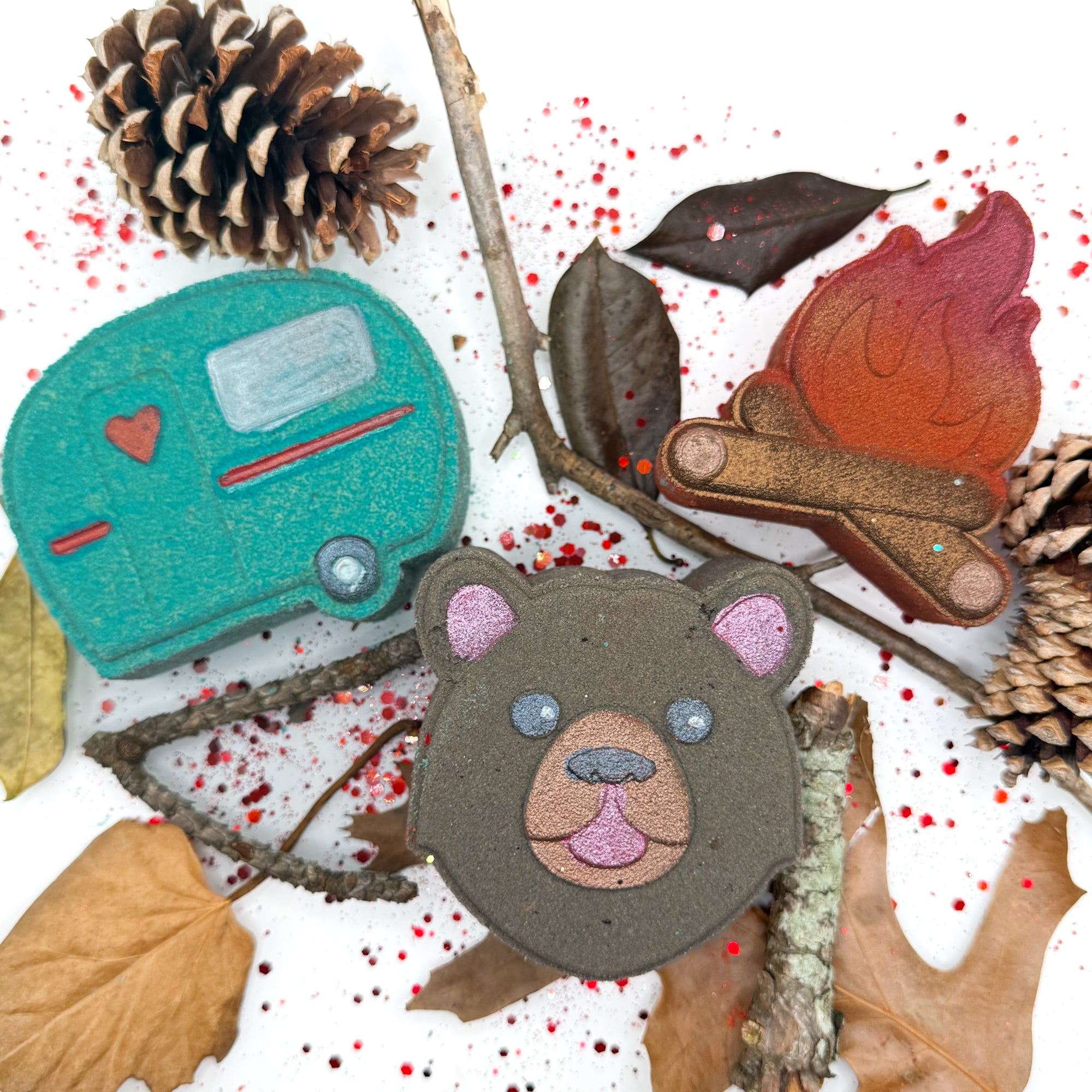 Three hand painted bath bombs (brown bear, teal camper and a campfire)with a camping theme on a white background. 