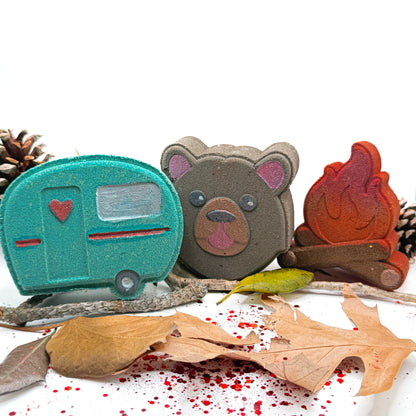 Three camping themed bath bombs shaped like a teal camper, brown bear, and orange campfire with decorative autumn leaves and pinecones on a white background.
