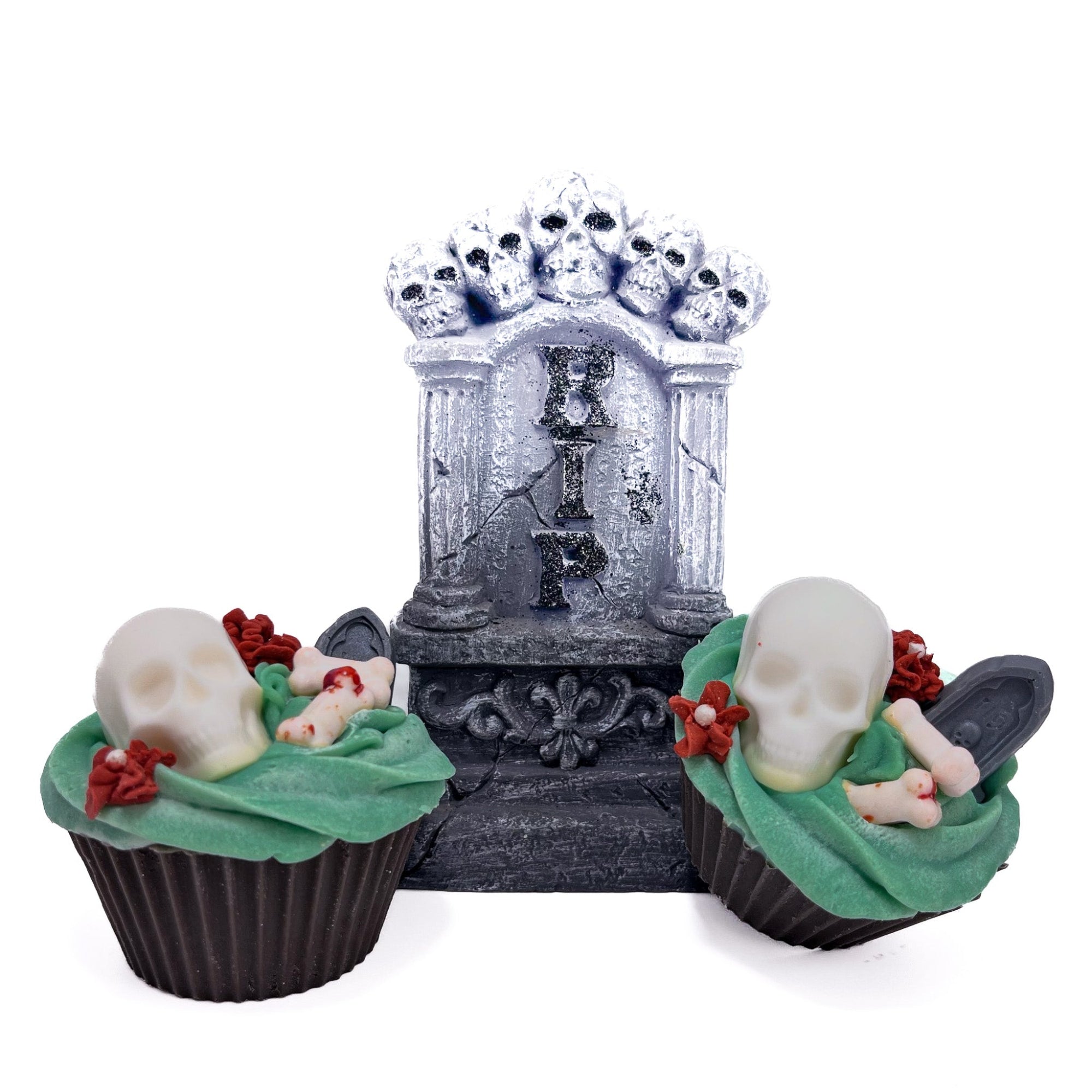 Two graveyard cupcake shaped bar soaps. Dirt brown cake, topped with green frosting, white skull and bones, grey tombstone, red flowers. Displayed with a gothic alter.