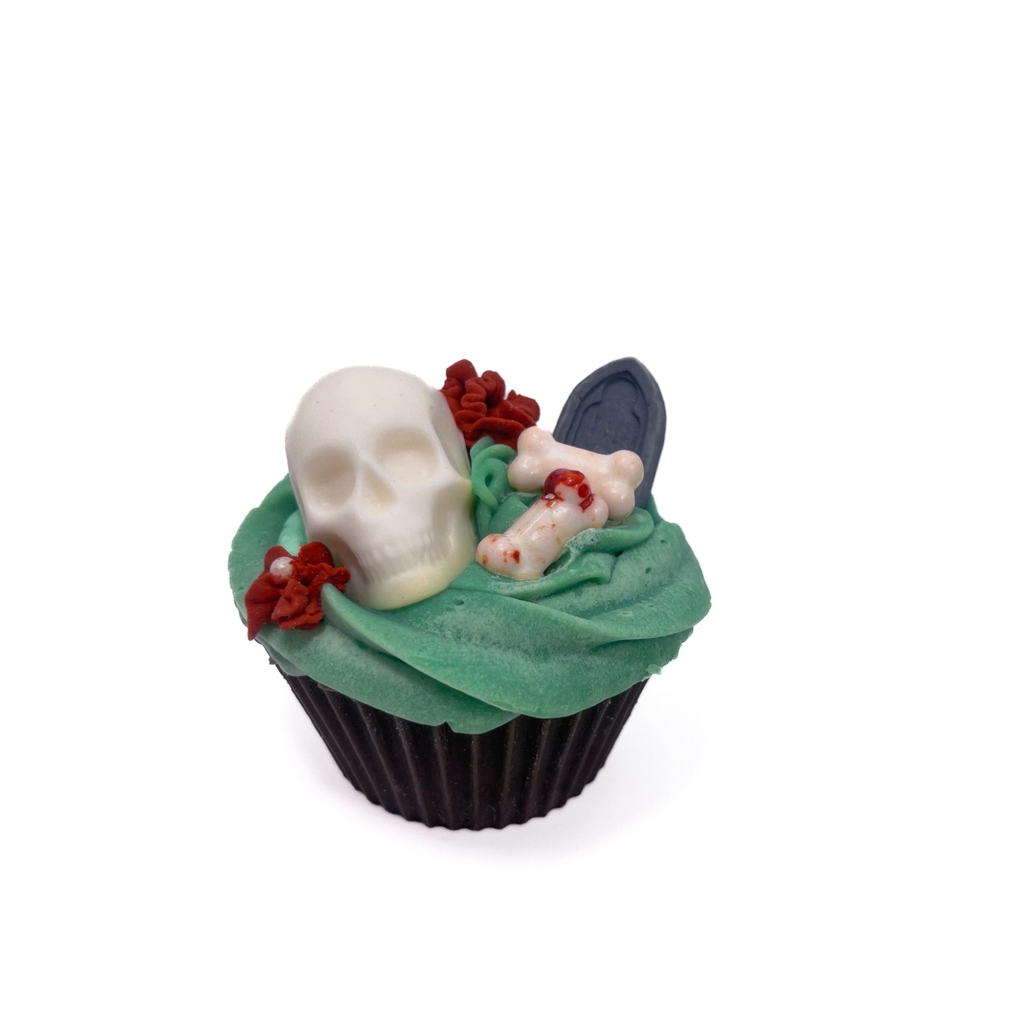 A graveyard cupcake shaped bar soap. Dirt brown cake, topped with green frosting, white skull and bones, grey tombstone, red flowers. 