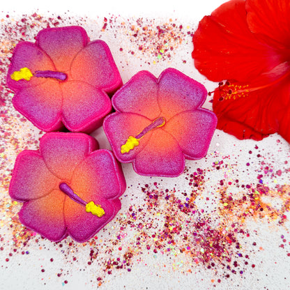 Three hibiscus shaped bath bombs with an airbrushed pink-to-orange gradient and yellow stamen details, arranged on a white surface with scattered glitter and a red hibiscus flower prop.