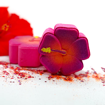 A close-up studio shot of hibiscus shaped bath bath bombs with a bright magenta-to-orange gradient and a small yellow center, arranged on a white surface with scattered red powder and a blurred red flower in the background.