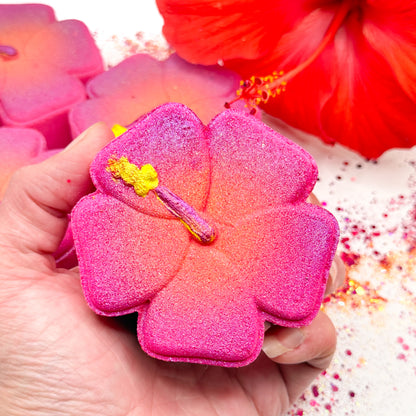 A hand holds a pink hibiscus shaped bath bomb with a hand painted yellow stamen detail, surrounded by other hibiscus bath bombs and a red hibiscus flower on a white background.