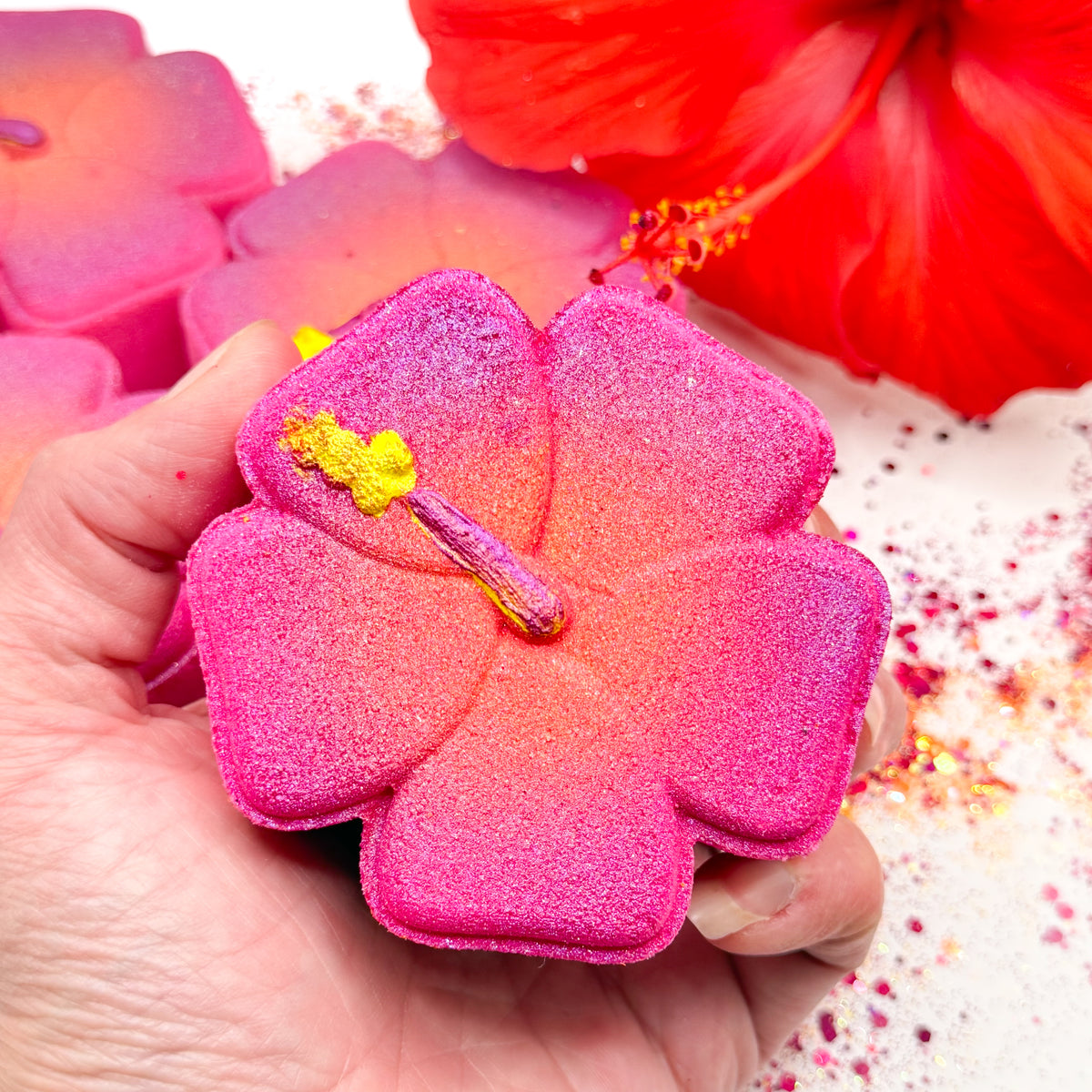 A hand holds a pink hibiscus shaped bath bomb with a hand painted yellow stamen detail, surrounded by other hibiscus bath bombs and a red hibiscus flower on a white background.