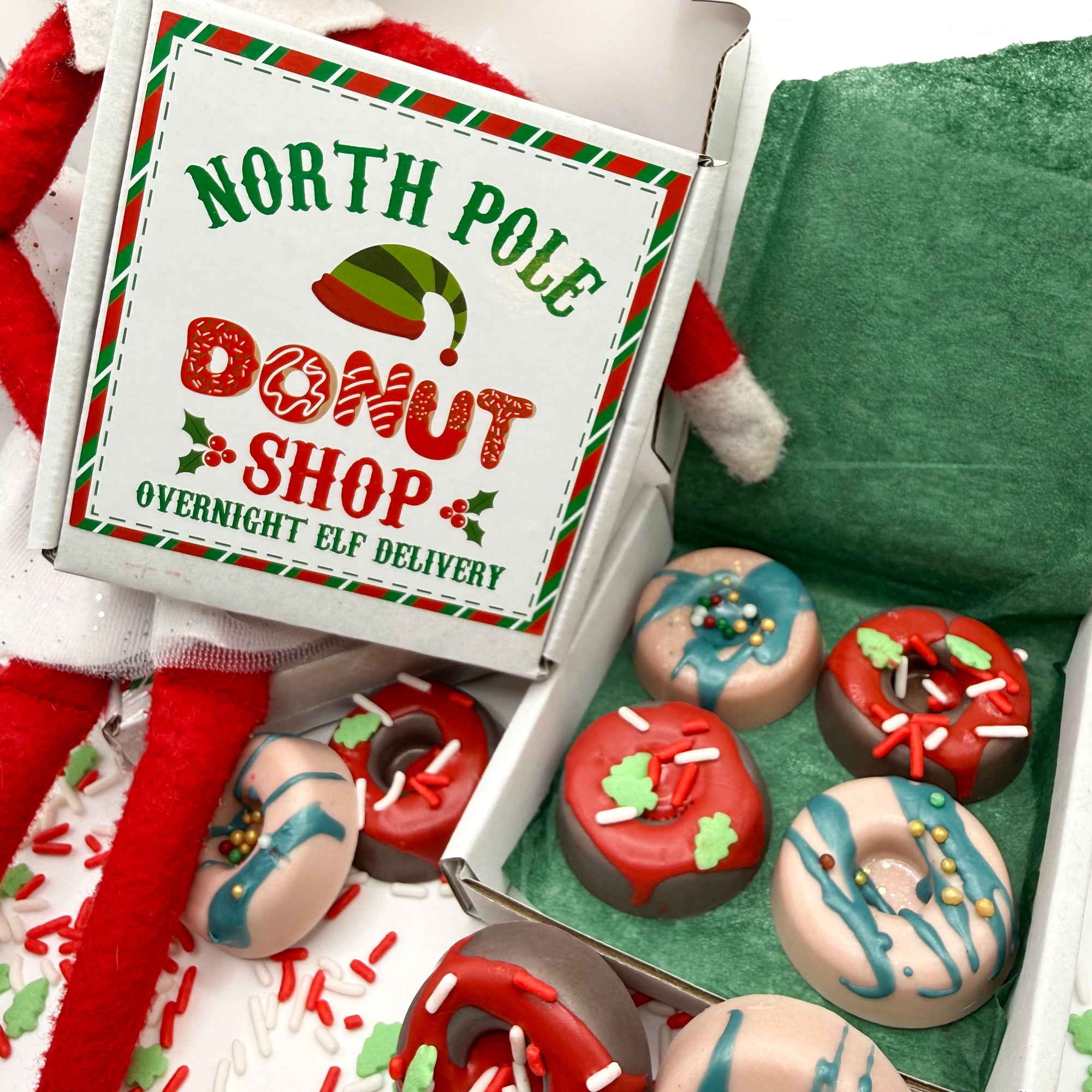 Box of 4 handmade red and green min donut soaps in a box that says 'North Pole Donut Shop'  Overnight elf delivery with Christmas themed decorations on box.