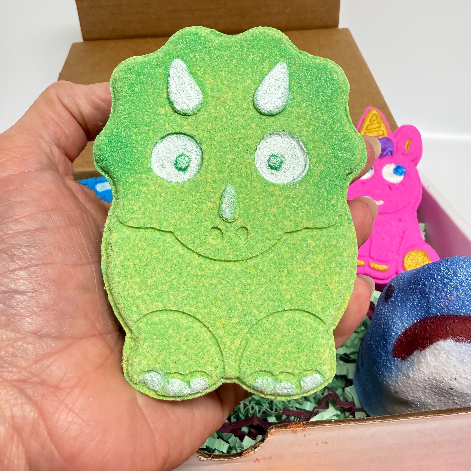 Green dinosaur bath bomb being held in a hand with other bath bombs in the back ground. 