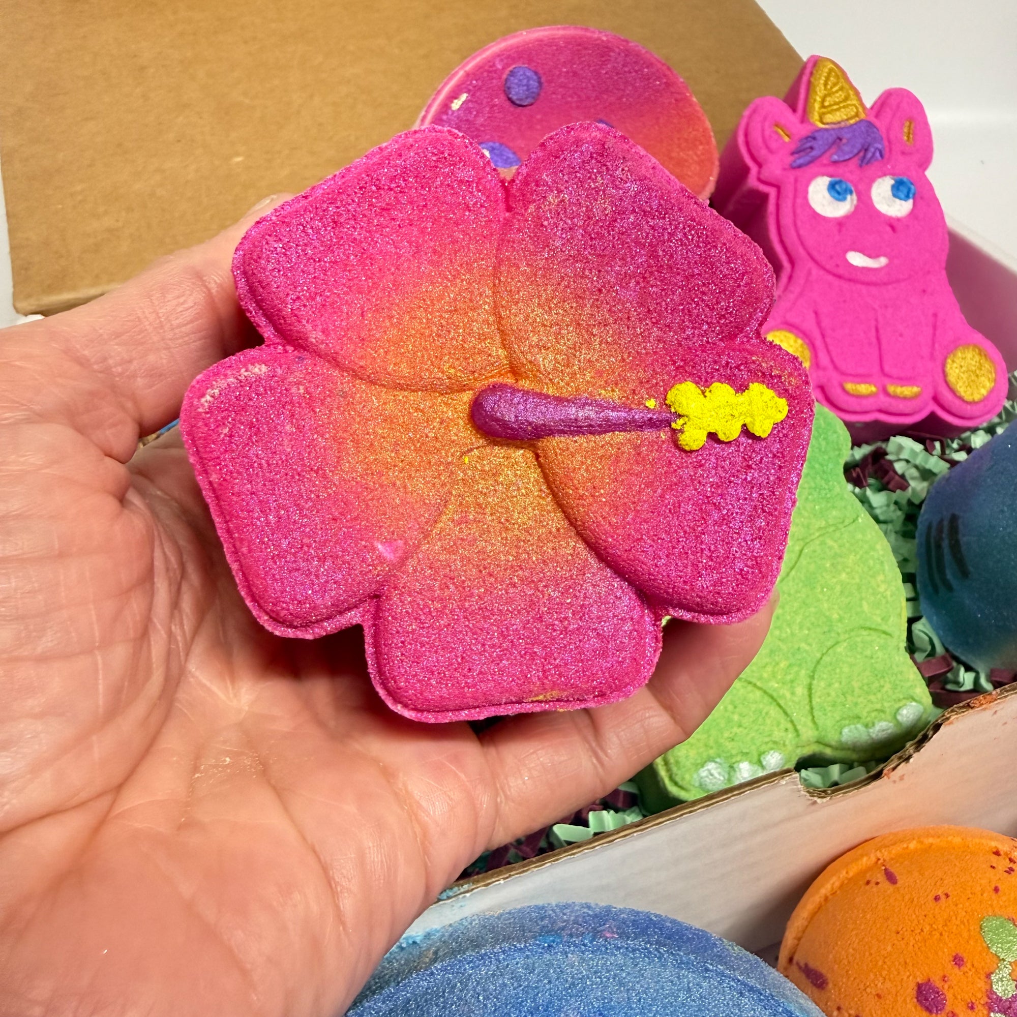 Pink hibiscus bath bomb held in a hand with other colorful bath bombs in the background. 
