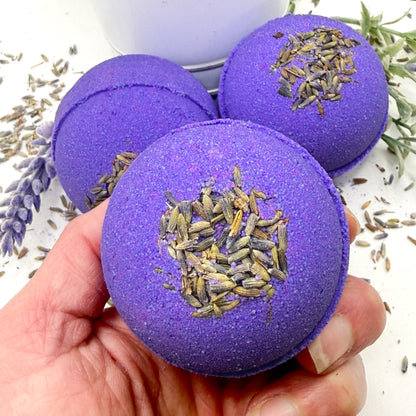 Purple lavender bath bomb with lavender buds held by a hand, surrounded by lavender flowers.