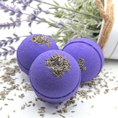 Three purple lavender bath bombs with lavender buds on a white surface with lavender flowers in the background.
