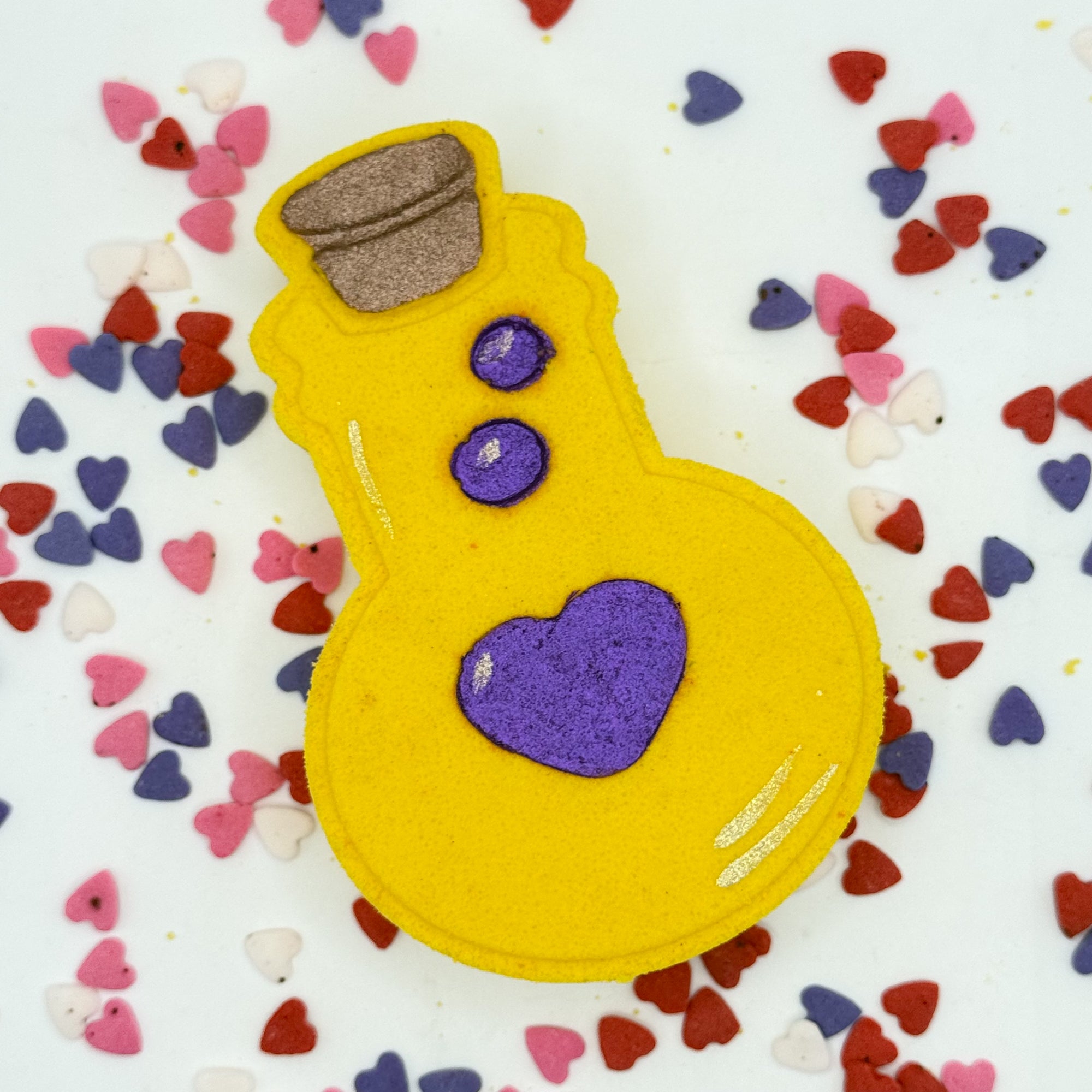 Handmade yellow bottle shaped bath bomb with purple hearts on a white background with colorful heart confetti